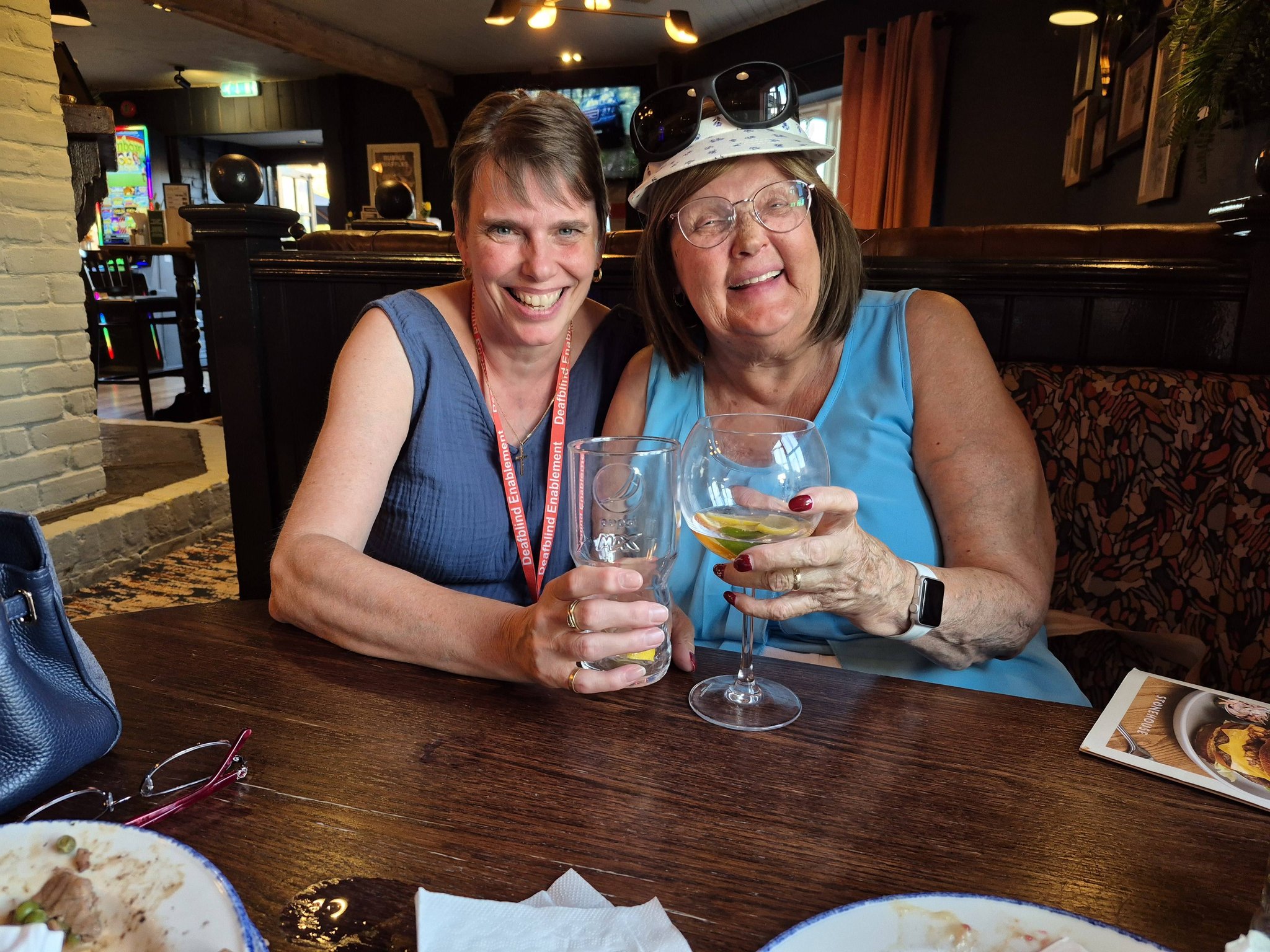 Two people sitting together in a pub booth, raising their drinks and laughing at the camera. One wears a red Deafblind Enablement lanyard.