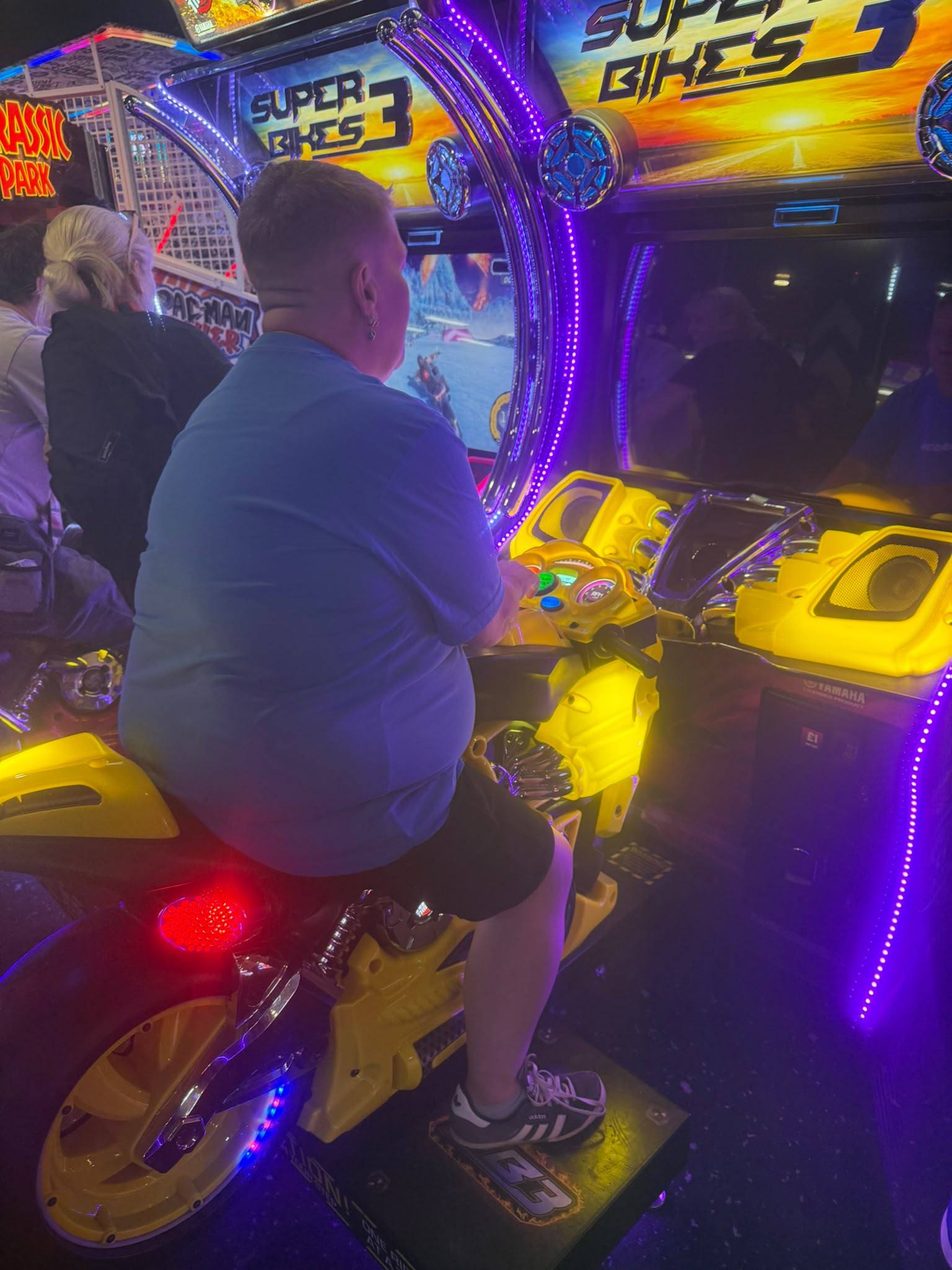 A service user riding the "Super Bikes" arcade motorbike at the bowling alley, leaning into the screen, surrounded by purple and yellow neon lights.