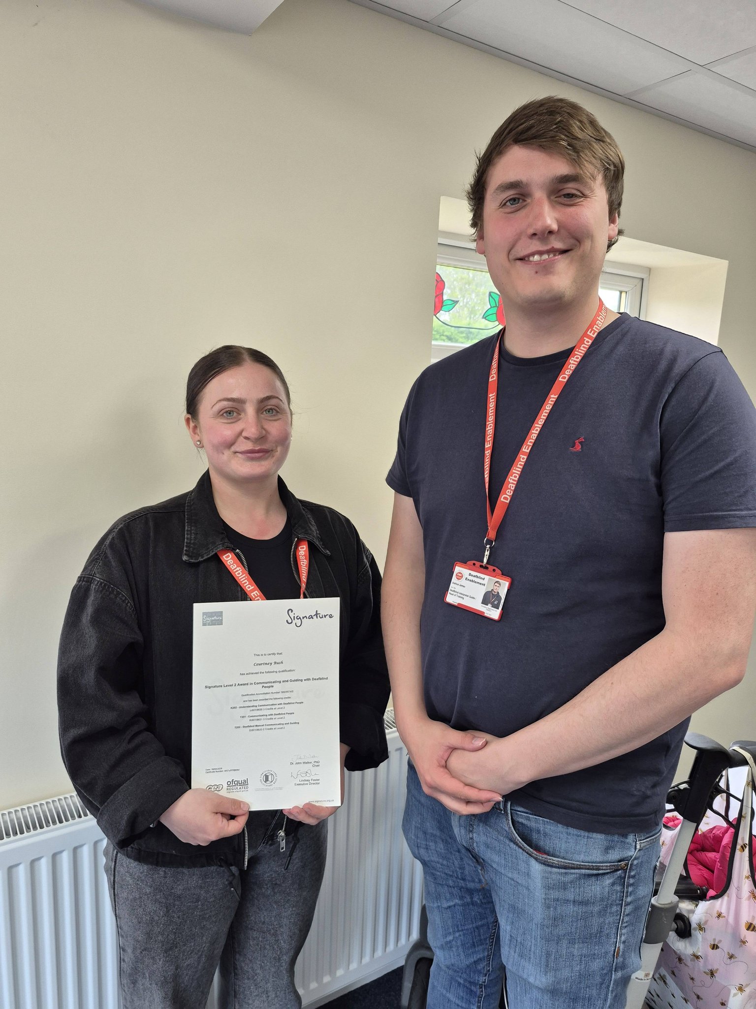Courtney holding her Signature Level 2 certificate, standing beside a colleague from the Deafblind Enablement training team who is wearing a red DBE lanyard.