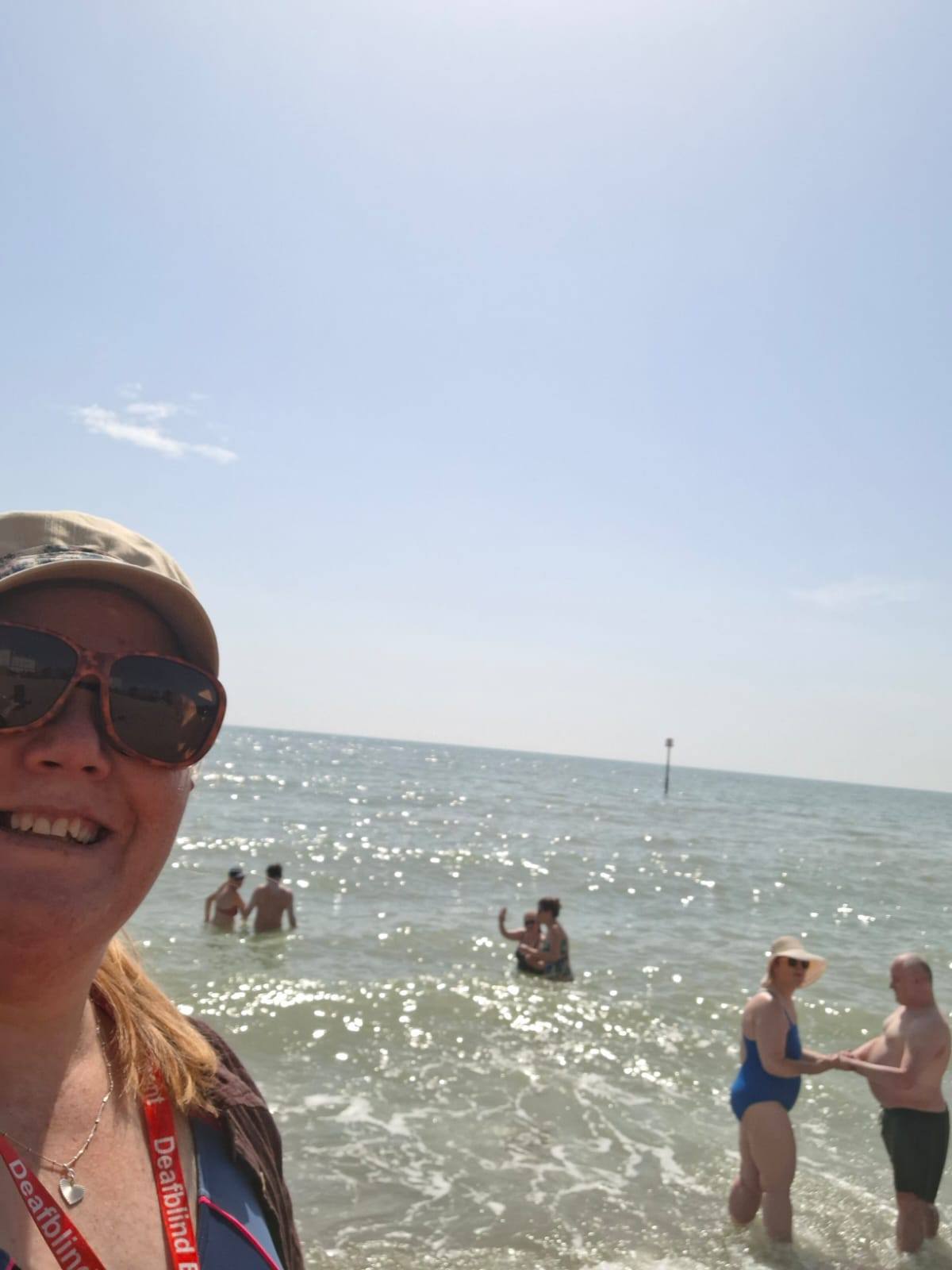 A Communicator-Guide wearing a Deafblind Enablement lanyard smiles in the foreground while service users and guides paddle and swim in the sea on a bright sunny day.
