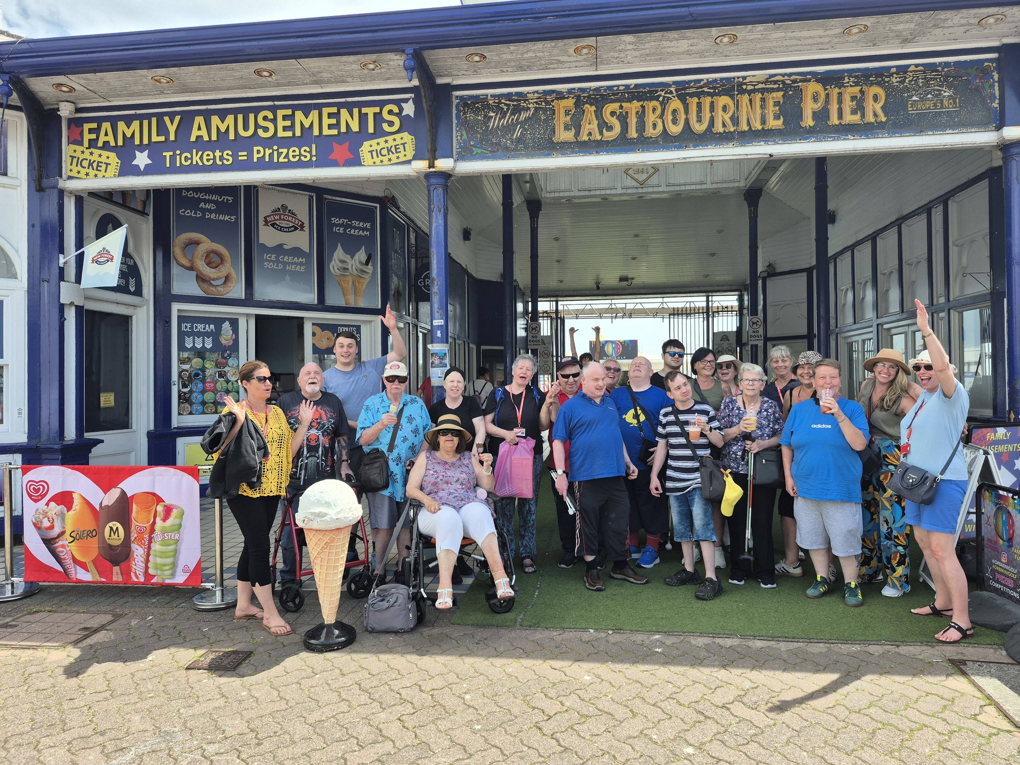 The whole Deafblind Enablement group standing together under the "Welcome to Eastbourne Pier" sign in bright sunshine, several people cheering with their arms in the air.