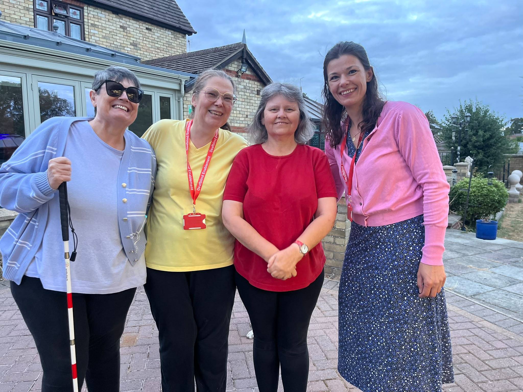 Four people standing together outside in the evening, smiling at the camera. One holds a white cane and two wear red Deafblind Enablement lanyards.