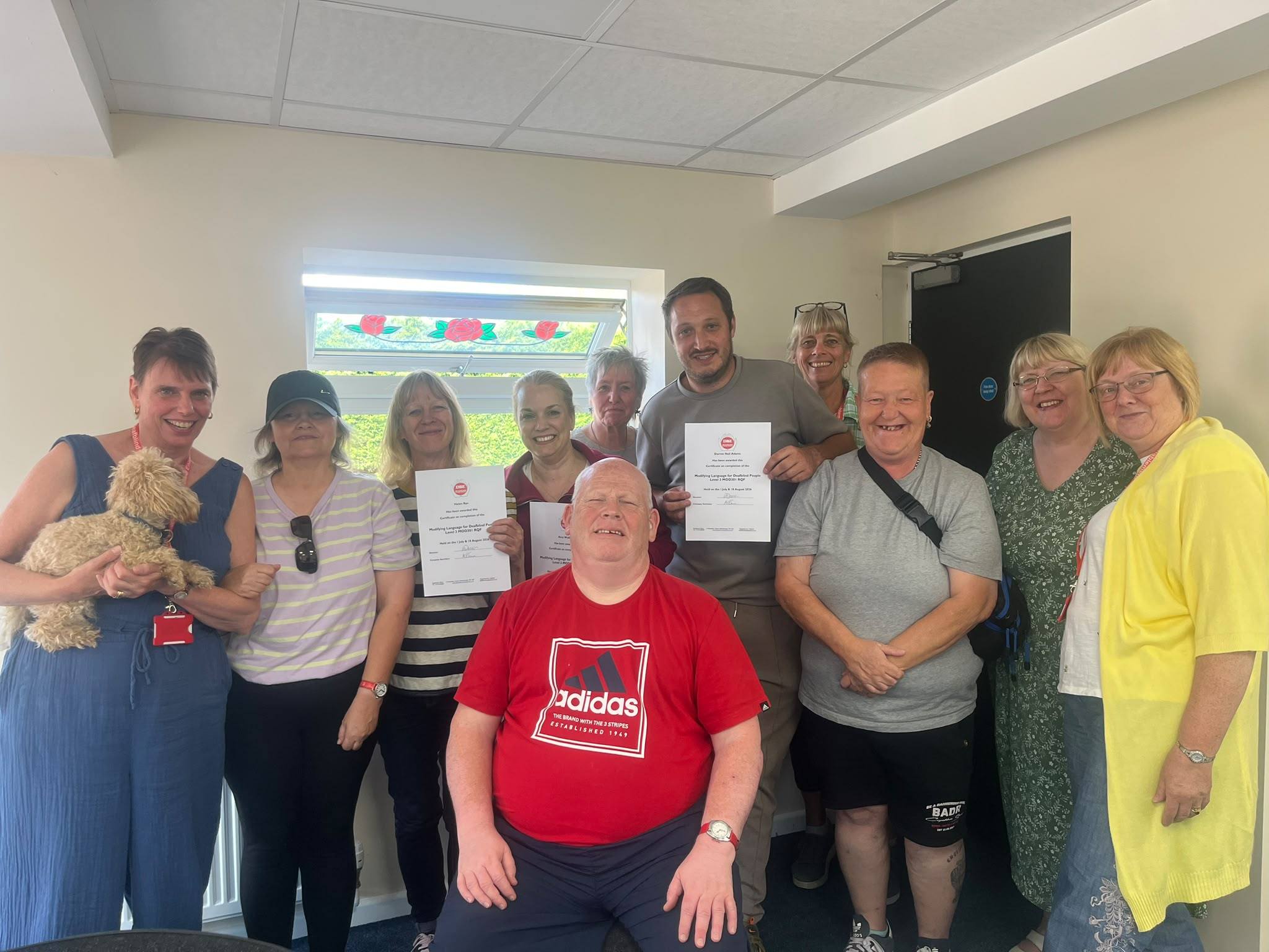 Eleven people standing together in a room at Deafblind Enablement, smiling at the camera. Three of them hold up their Modifying Language for Deafblind People certificates, and the woman on the left is holding a small cream-coloured dog.