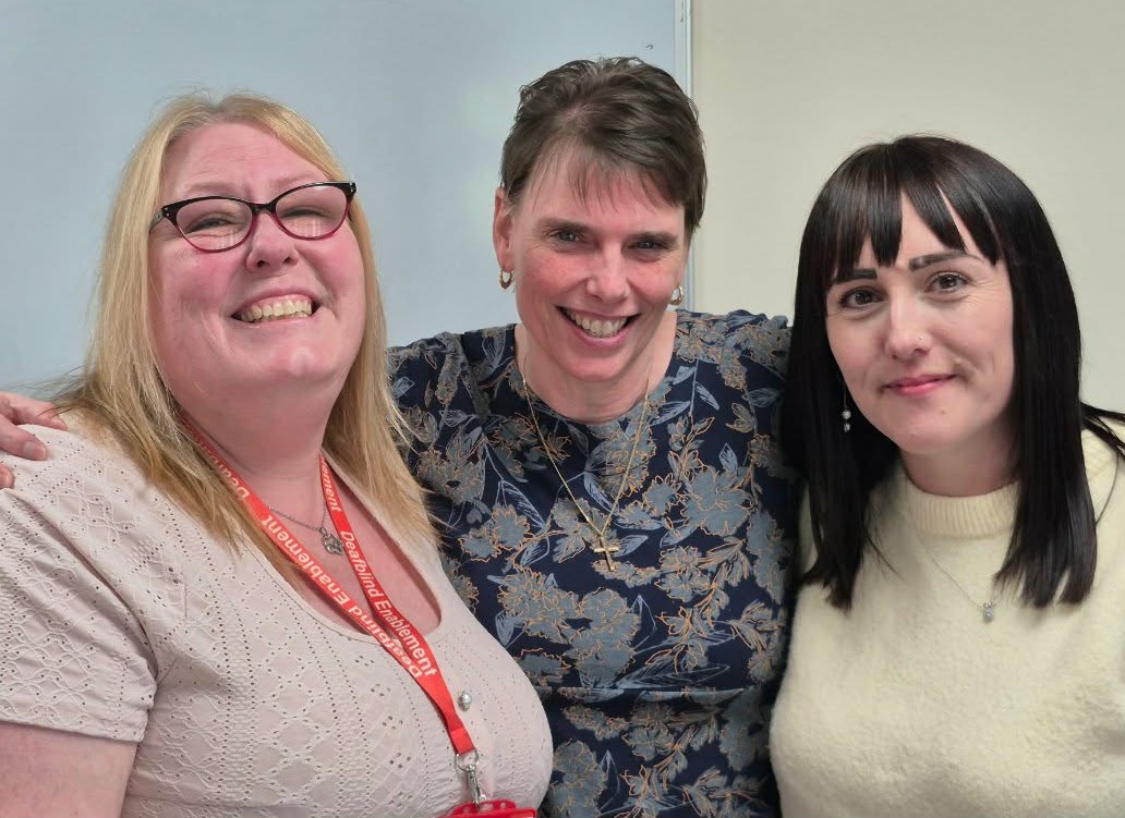 Three members of the Deafblind Enablement team standing together and smiling at Head Office in Peterborough. One wears a red Deafblind Enablement lanyard.